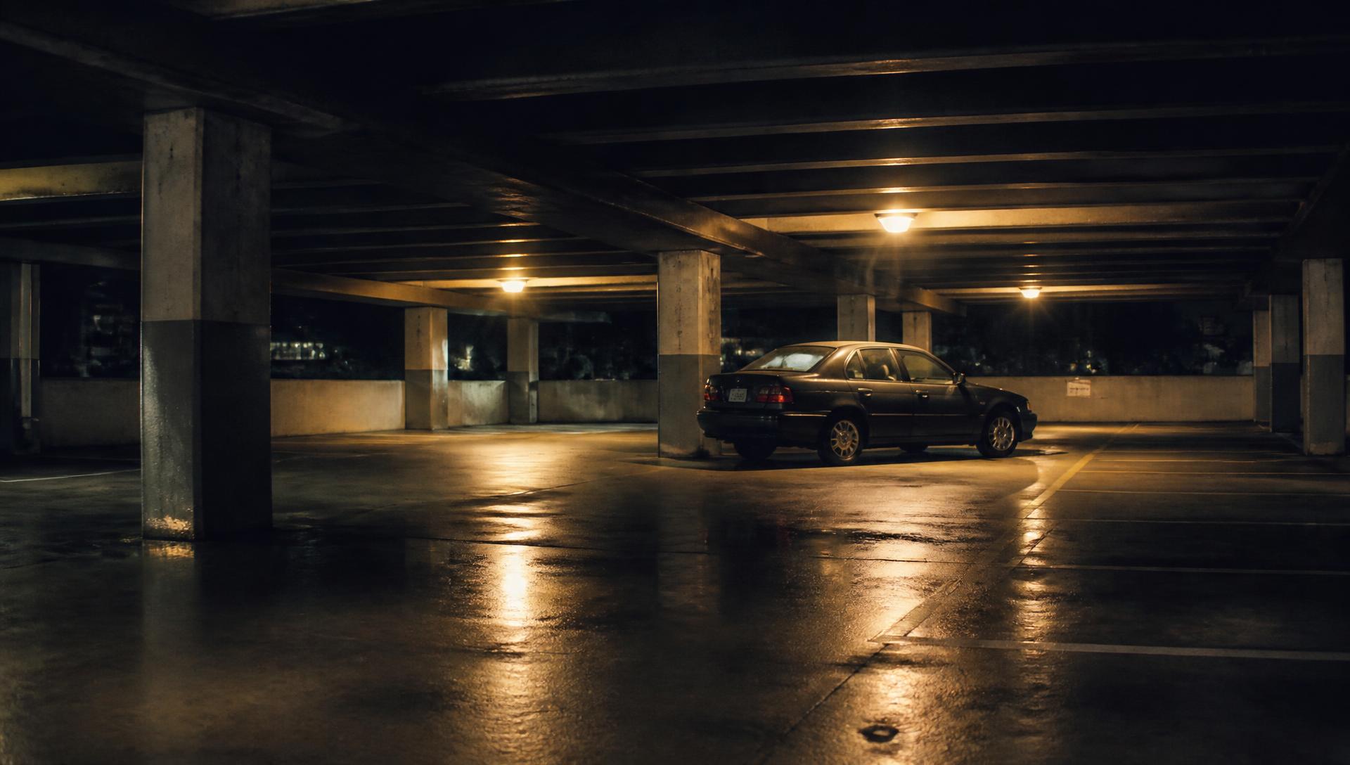 An empty concrete parking garage at night with one car parked under an amber lamp